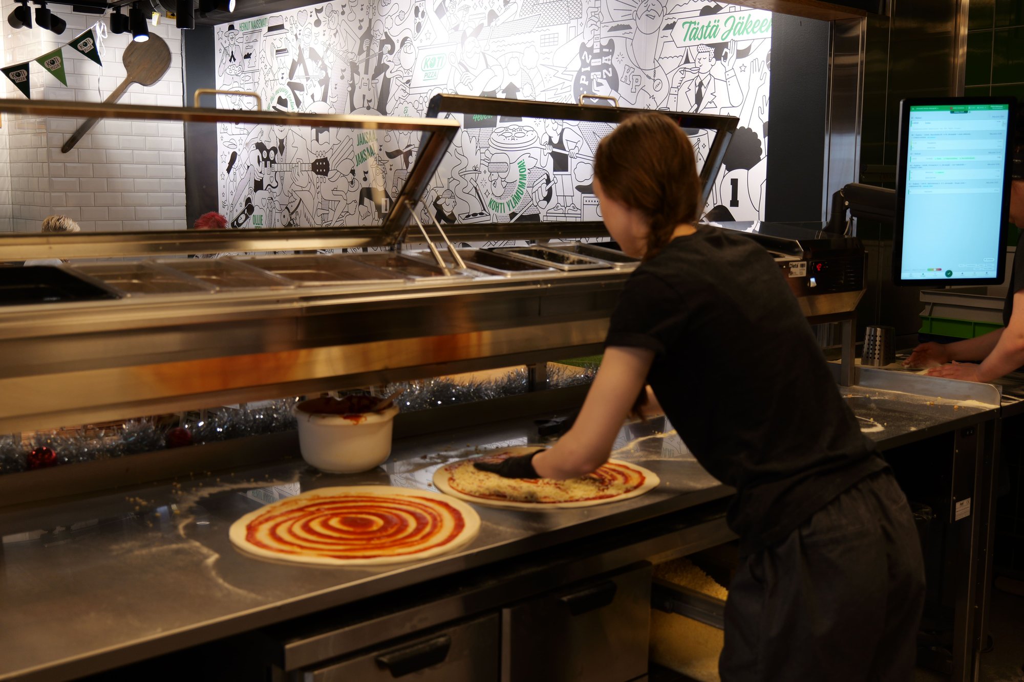 Kotipizza employee preparing pizzas with a Kuori kitchen display nearby. Kotipizza employee preparing pizzas with a Kuori kitchen display nearby.